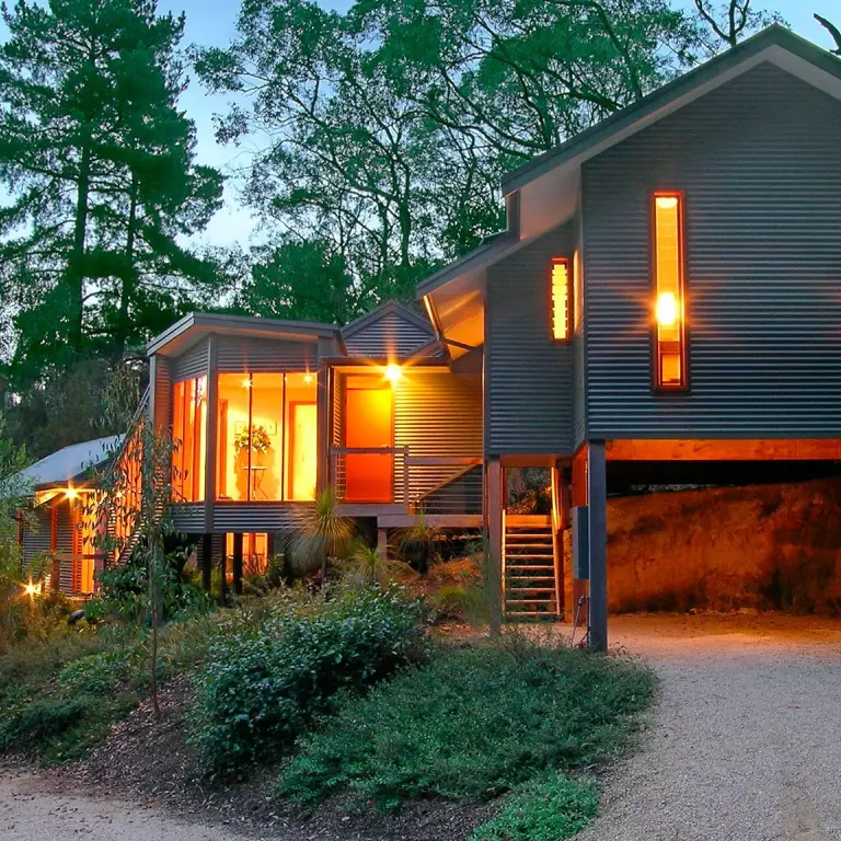 Zeitgenössisches Cottage auf Stelzen mit Wellblechverkleidung und warm leuchtendem Innenraum, eingebettet zwischen hohen Bäumen im australischen Busch in der Abenddämmerung