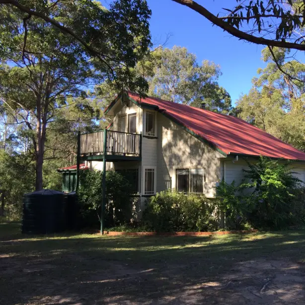 Freestanding two-storey cottage with red tin roof and balcony, surrounded by eucalyptus trees and open lawn in the Australian bush on a sunny day
