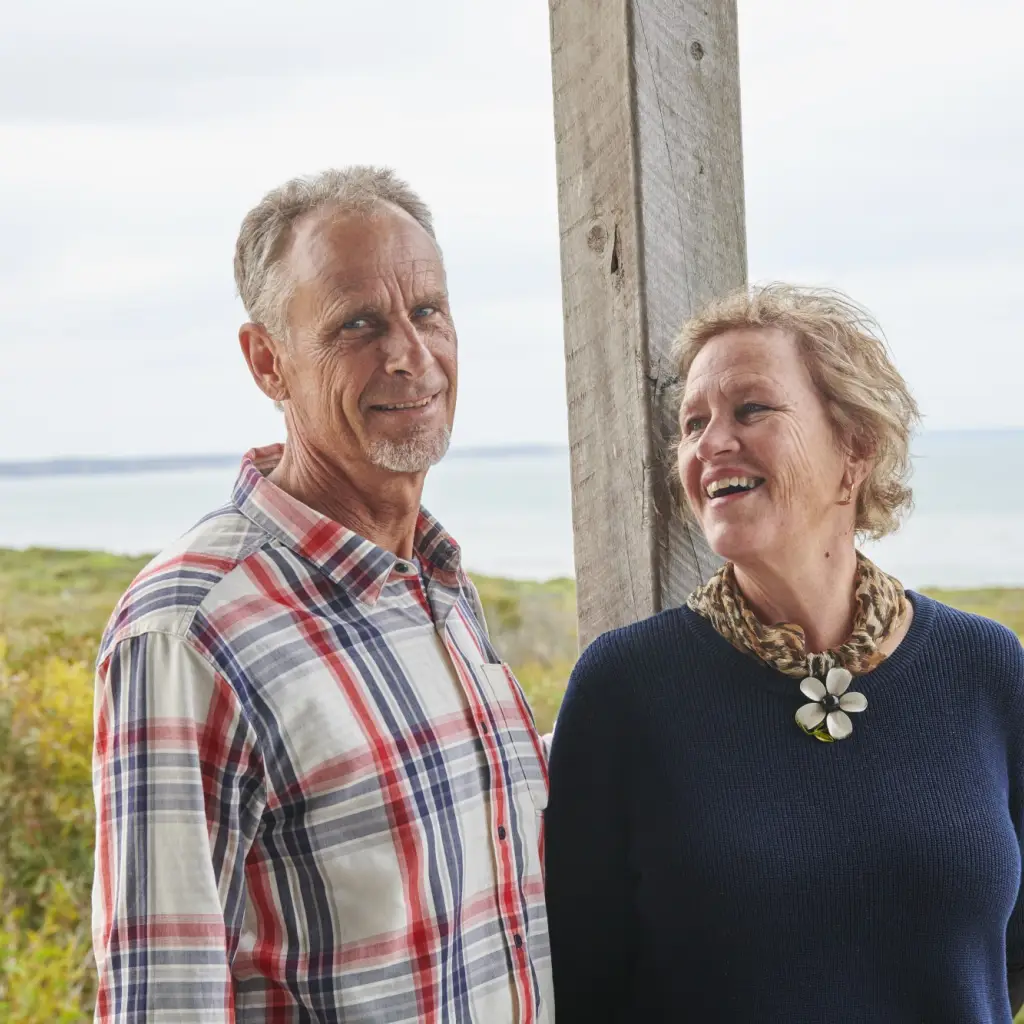 A couple stand against a wooden beam smiling with bush and ocean in the background