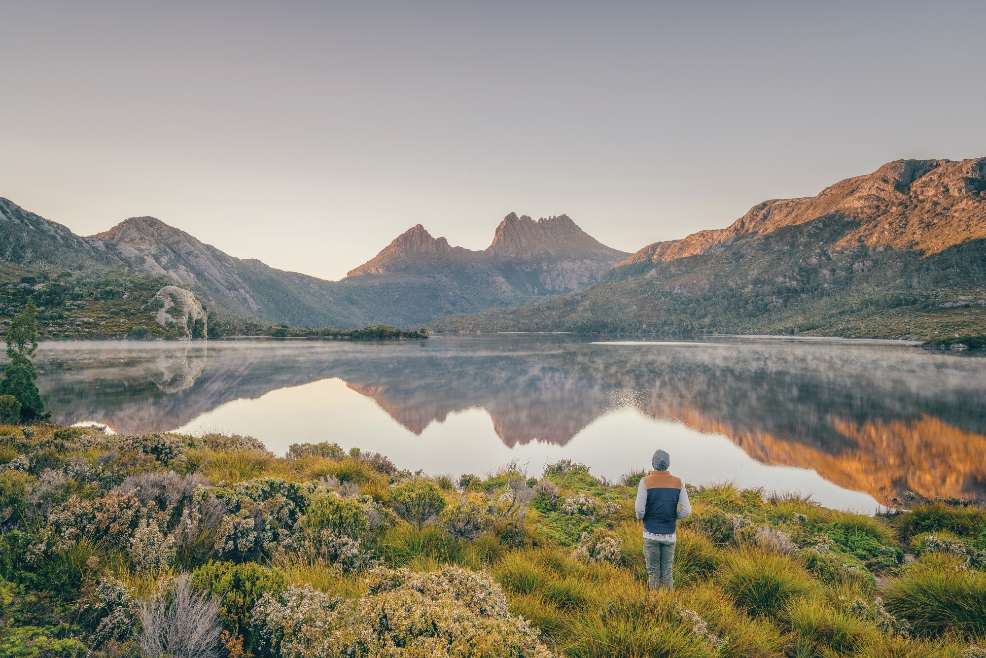 Der Cradle Mountain Australia spiegelt sich bei Sonnenaufgang in einem ruhigen See, an dessen Ufer ein Reisender mit Blick auf die Bergkette steht.