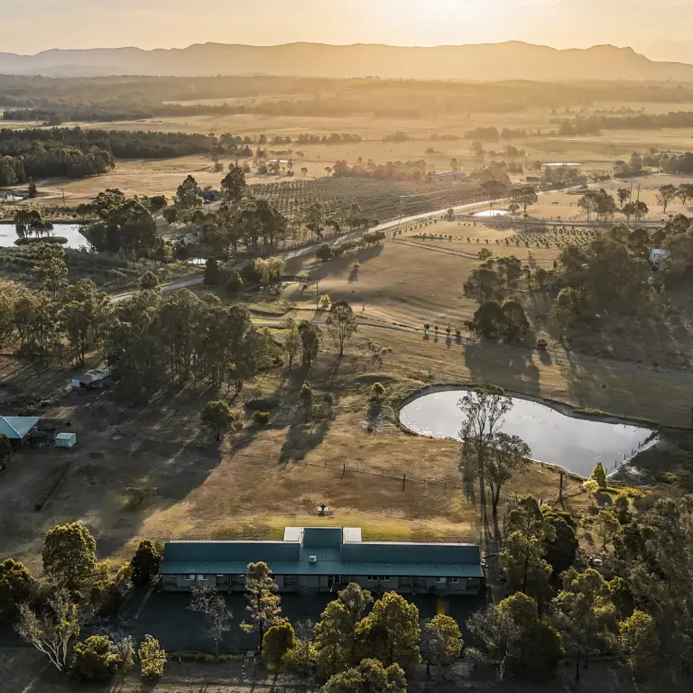 Luchtfoto van een boerderijaccommodatie in Australië tijdens het gouden uur, met een landelijke lodge omgeven door glooiende weilanden, boerderijdammen, boomgaarden en eucalyptusbomen met bergen in de mistige achtergrond.