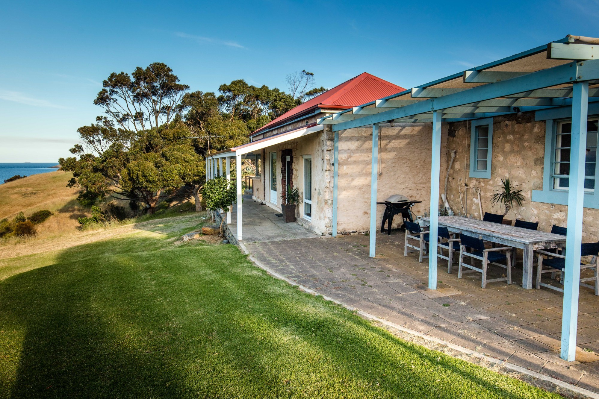 Historisches Steinferienhaus in Australien mit überdachter Außenterrasse und Grill, auf einem Klippenrasen mit weitem Blick über das Meer an einem klaren sonnigen Tag