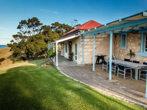 Historisches Steinferienhaus in Australien mit überdachter Außenterrasse und Grill, auf einem Klippenrasen mit weitem Blick über das Meer an einem klaren sonnigen Tag