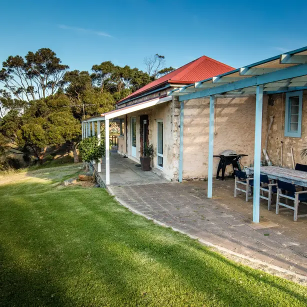 Historic stone holiday house in Australia with a covered outdoor dining terrace and barbecue, set on a clifftop lawn with sweeping views over the ocean on a clear sunny day