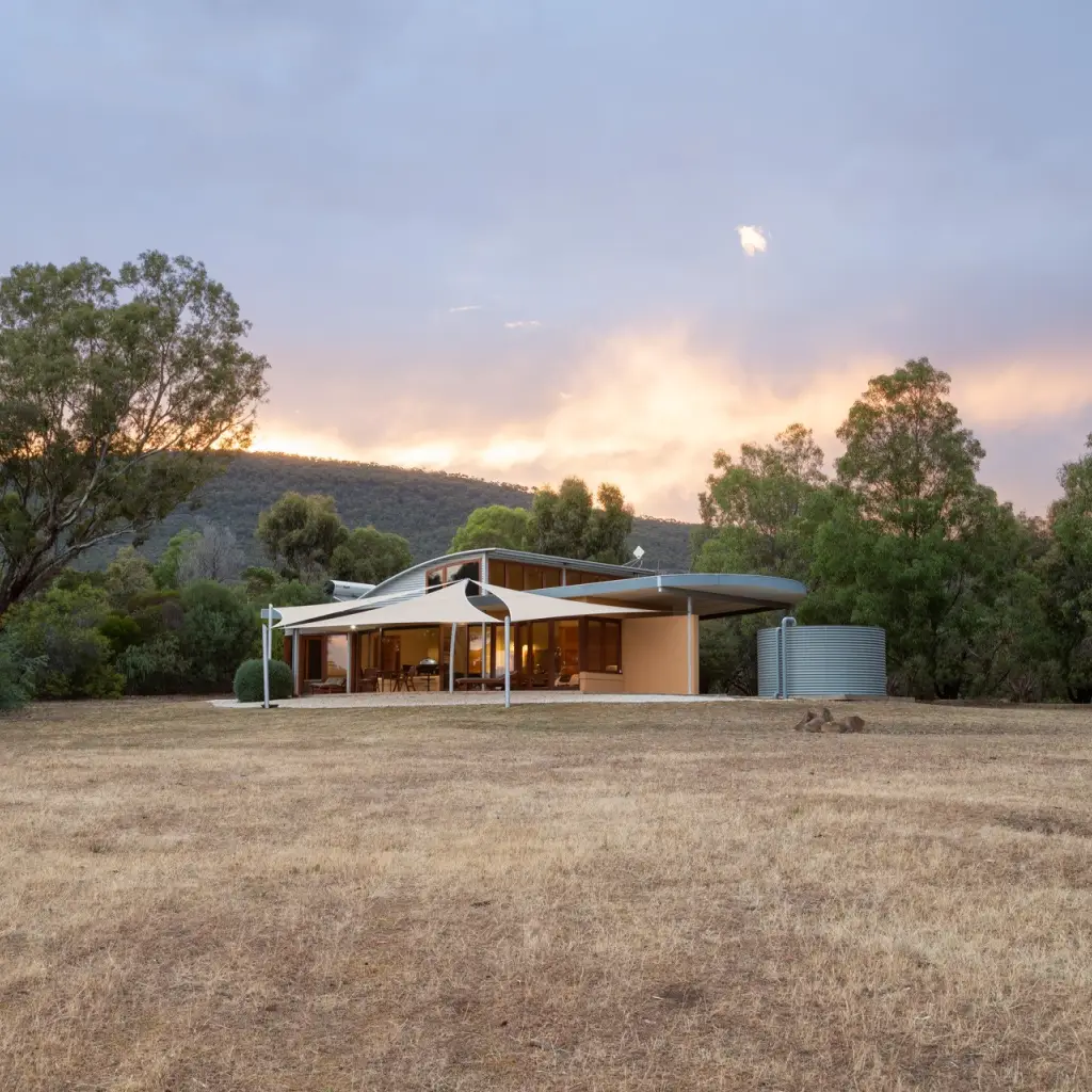 Zeitgenössisches, von einem Architekten entworfenes Ferienhaus mit geschwungenem Dach und Sonnensegeln, in einer offenen Grassavanne umgeben von Eukalyptusbäumen und Hügeln bei Sonnenuntergang im ländlichen Australien