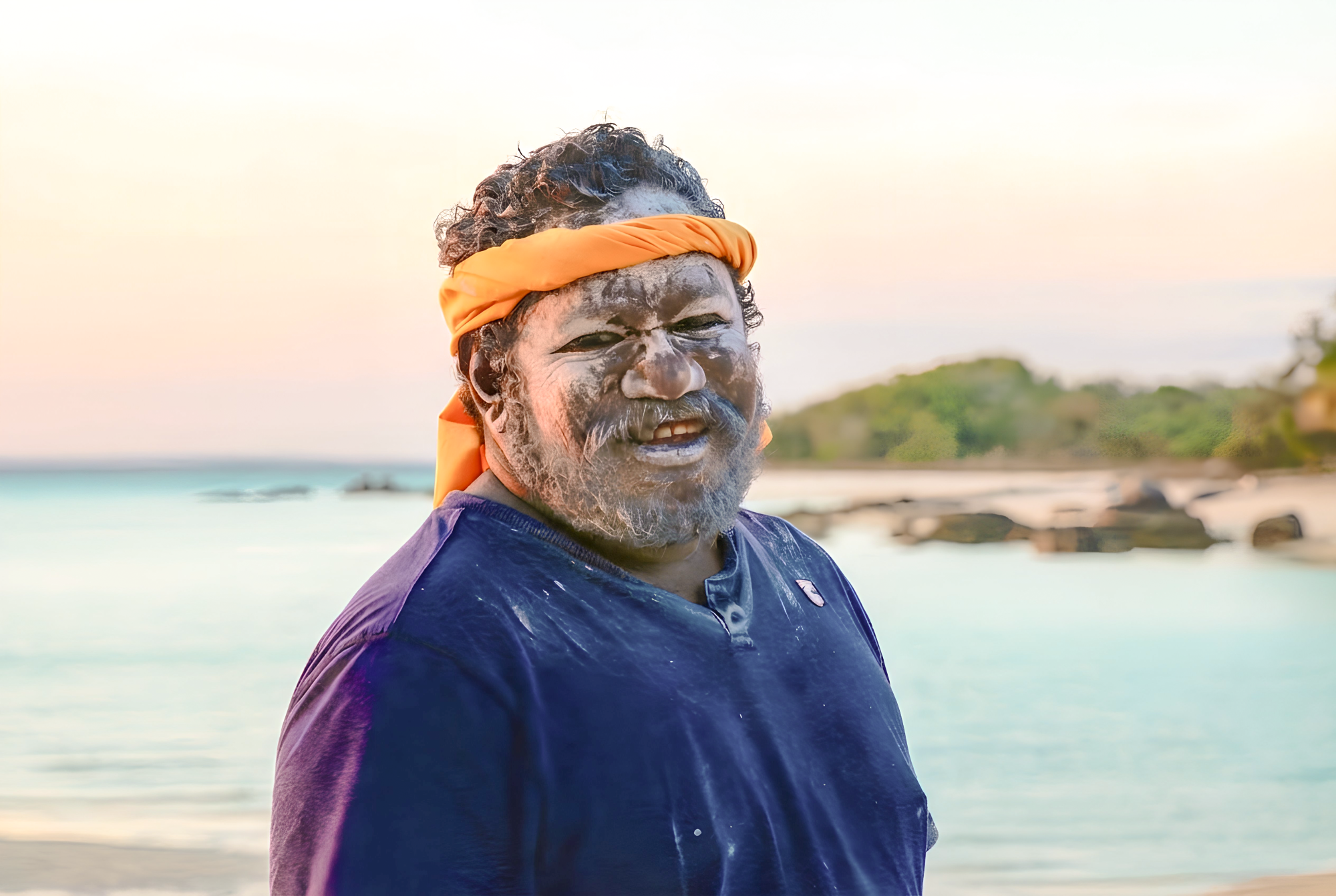 Smiling indigenous Australian man with traditional white face paint and an orange headband on a tropical beach at sunset, Australia