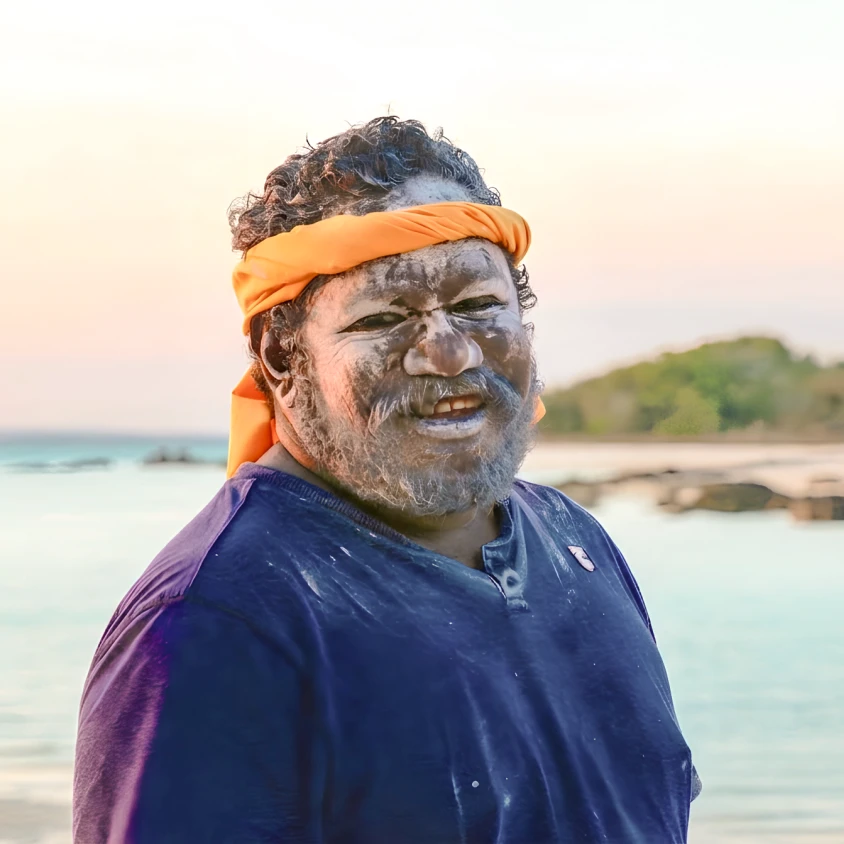 Een glimlachende Australische inheemse man met traditionele witte gezichtsverf en een oranje hoofdband op een tropisch strand bij zonsondergang, Australië