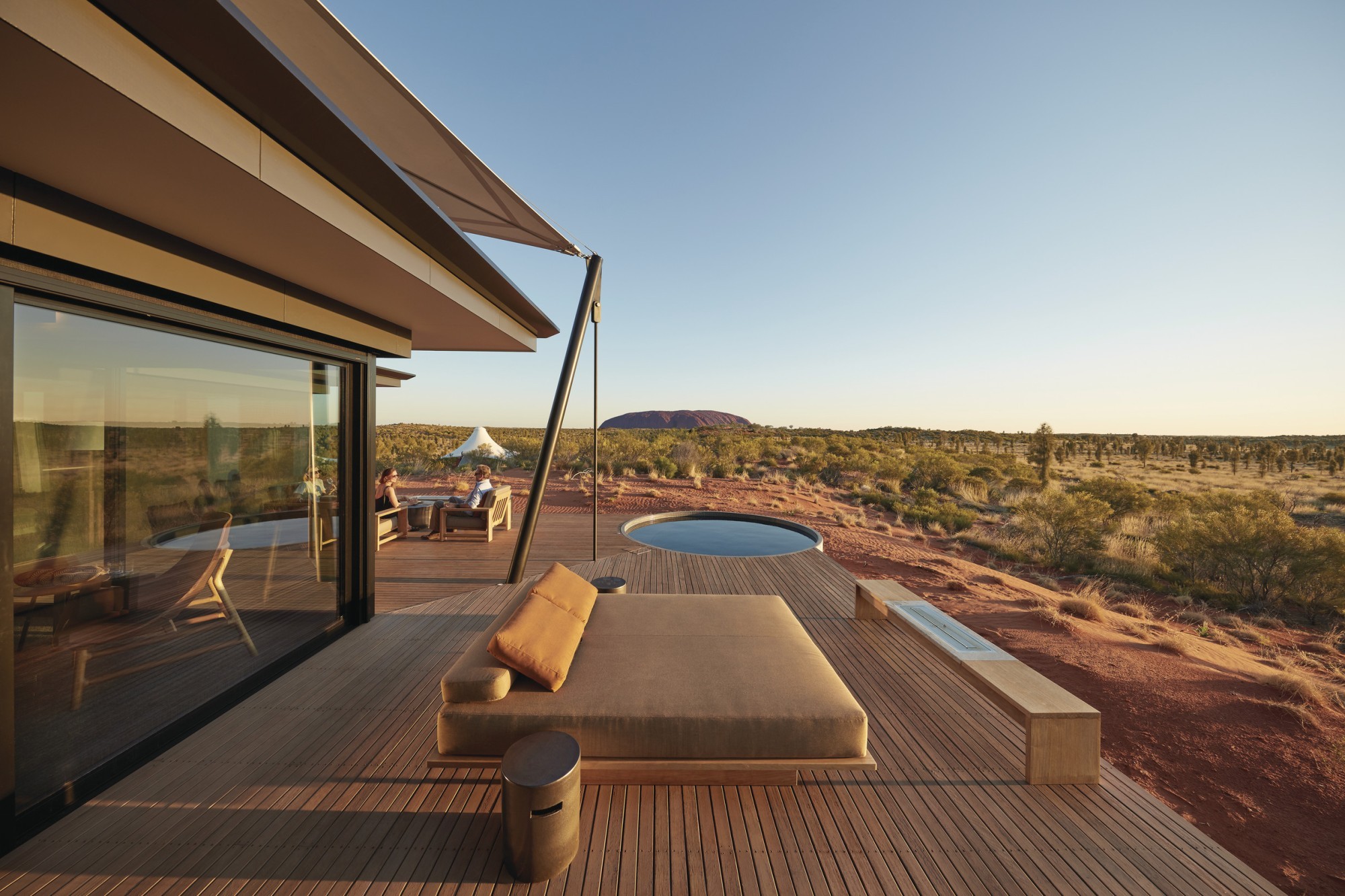 Luxuriöse Lodge-Terrasse mit Tagesbett und Tauchbecken mit Blick auf die rote Wüstenlandschaft von Uluru im Northern Territory Australiens, mit einem Paar im Hintergrund, das in der goldenen Stunde sitzt.