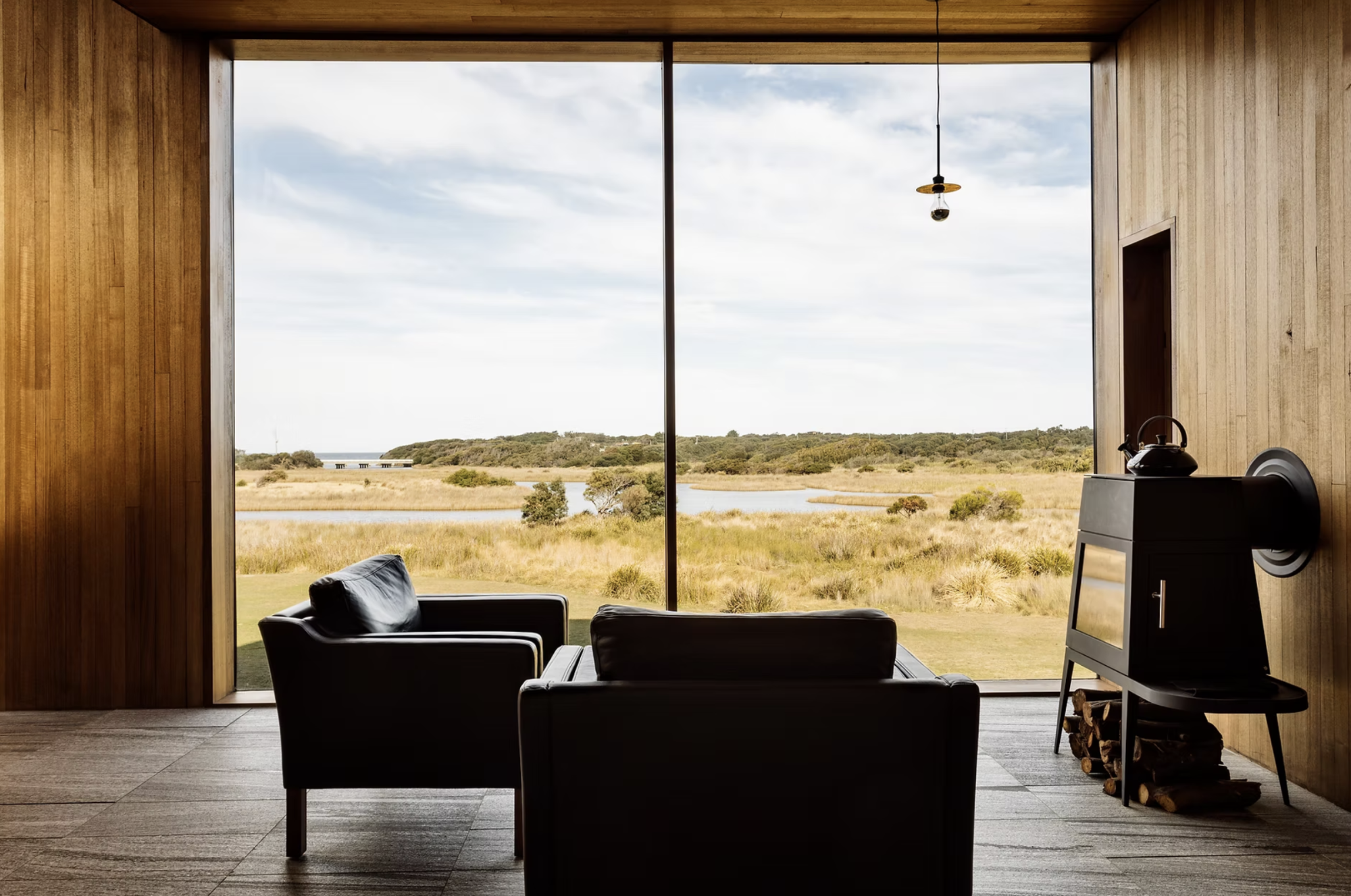 Two armchairs facing a large picture window overlooking open grassland, water and distant bushland, with a wood stove on the right.