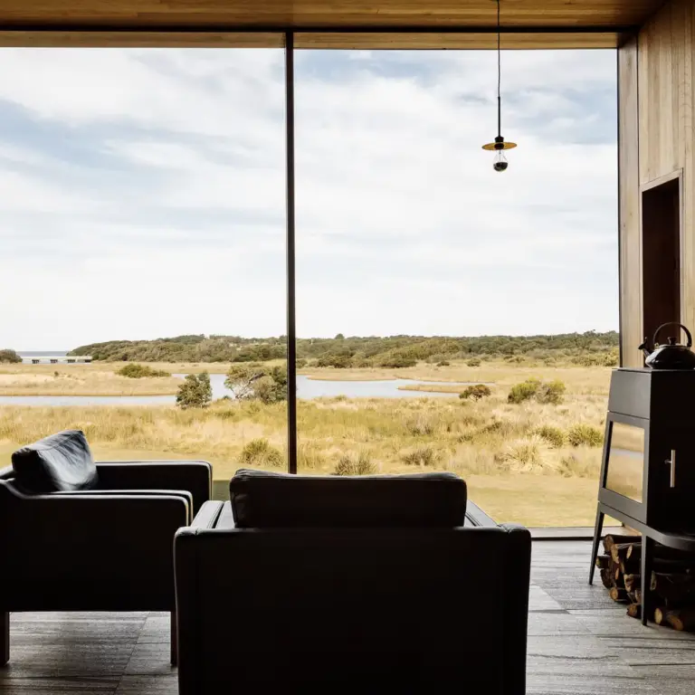 Two armchairs facing a large picture window overlooking open grassland, water and distant bushland, with a wood stove on the right.