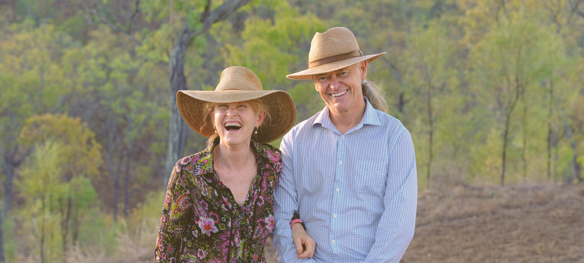 Smiling couple wearing wide-brimmed hats in the Australian outback of Queensland, local farmstay hosts welcoming guests in a rural bush setting