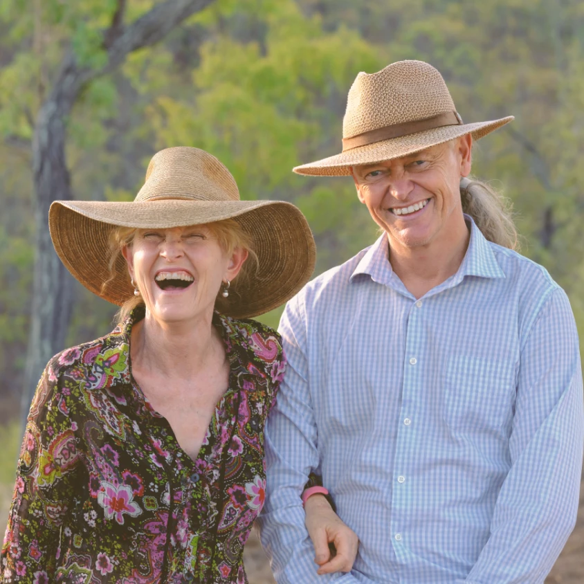 Een glimlachend stel met hoeden met brede randen in de Australische outback van Queensland; de gastheren van een lokale boerderij verwelkomen hun gasten in een landelijke bush-omgeving