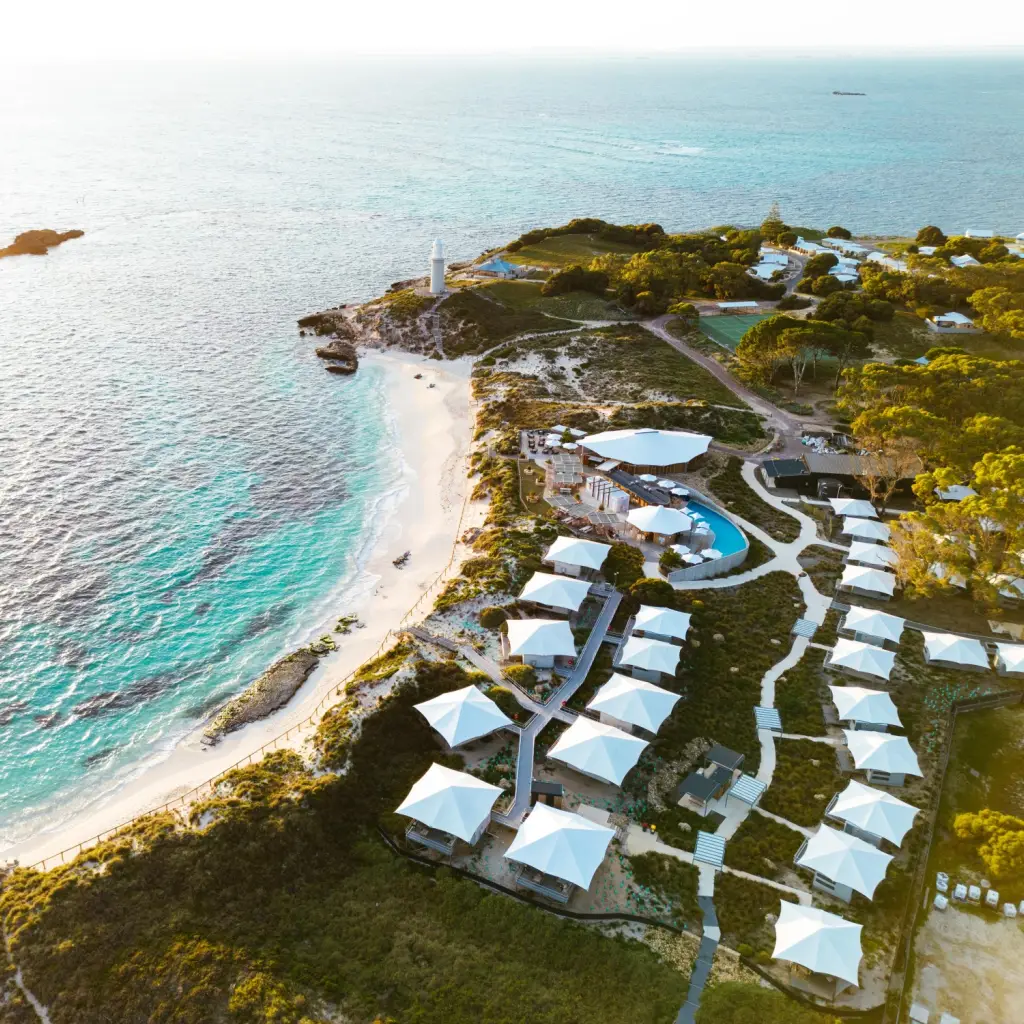 Luftaufnahme eines Strandresorts auf Rottnest Island, Australien, mit weißen Glamping-Zelten entlang einer türkisfarbenen Küste mit weißen Sandstränden und einem Leuchtturm bei Sonnenuntergang