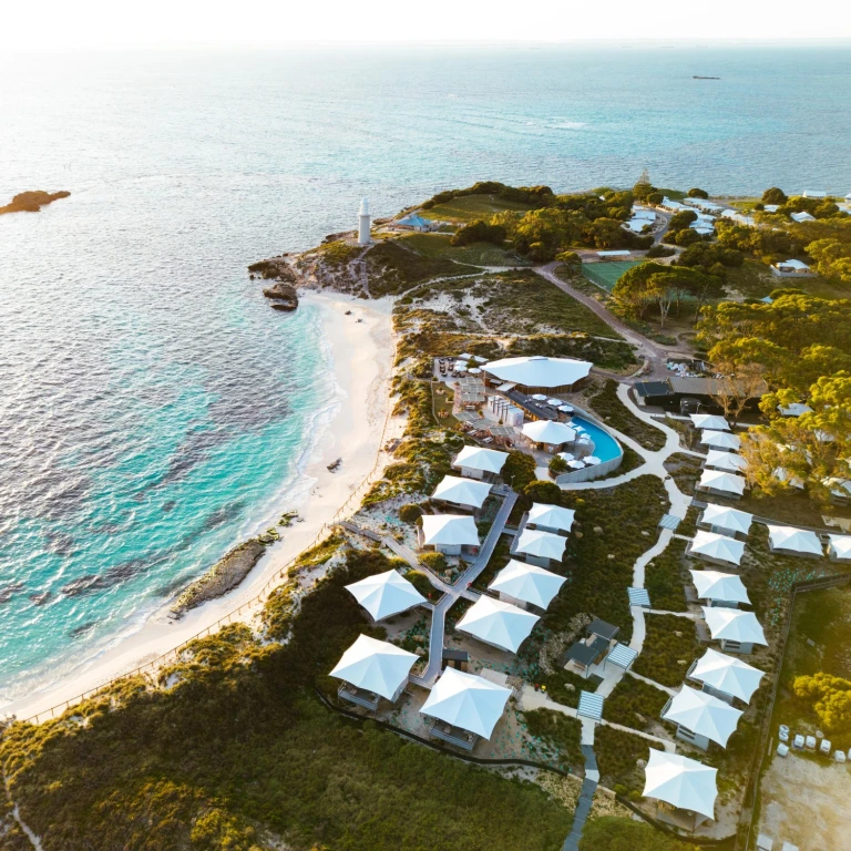 Luftaufnahme eines Strandresorts auf Rottnest Island, Australien, mit weißen Glamping-Zelten entlang einer türkisfarbenen Küste mit weißen Sandstränden und einem Leuchtturm bei Sonnenuntergang