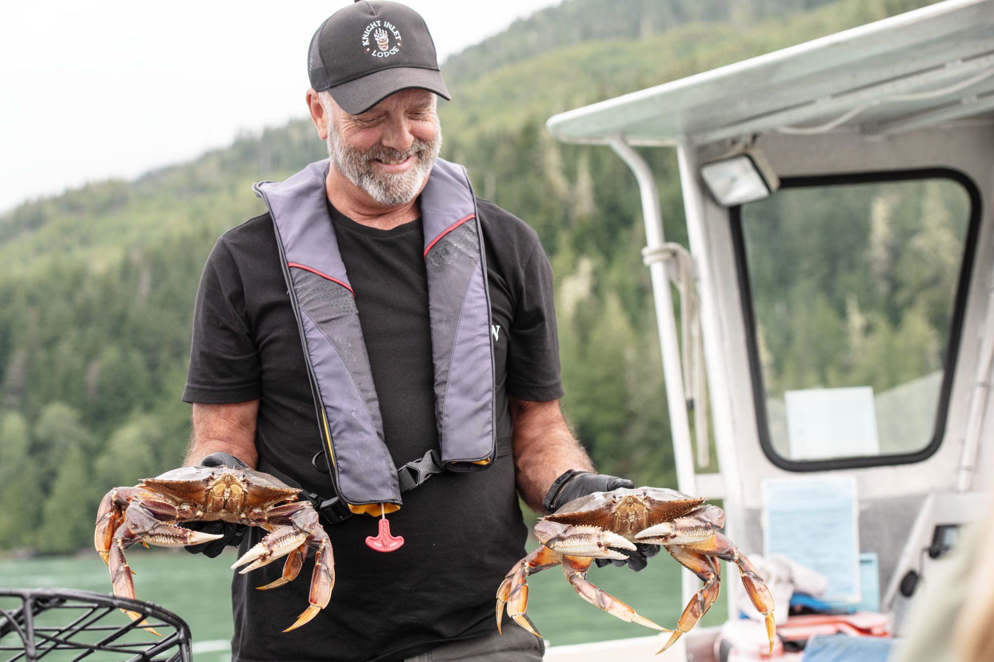 Ein lächelnder Guide der Knight Inlet Lodge hält zwei frisch gefangene Dungeness-Krabben auf einem Boot in die Höhe, mit bewaldeten Fjordhängen im Hintergrund, am Knight Inlet, British Columbia, Kanada.