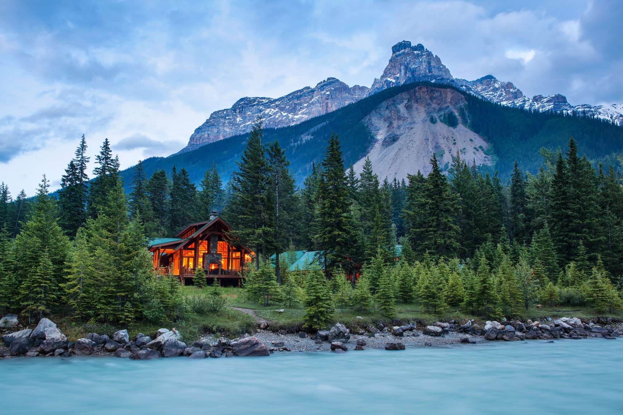 Warm-lit timber mountain lodge nestled among spruce trees on the bank of a glacier-fed turquoise river, with dramatic rocky peaks rising behind in Yoho National Park, British Columbia, Canada