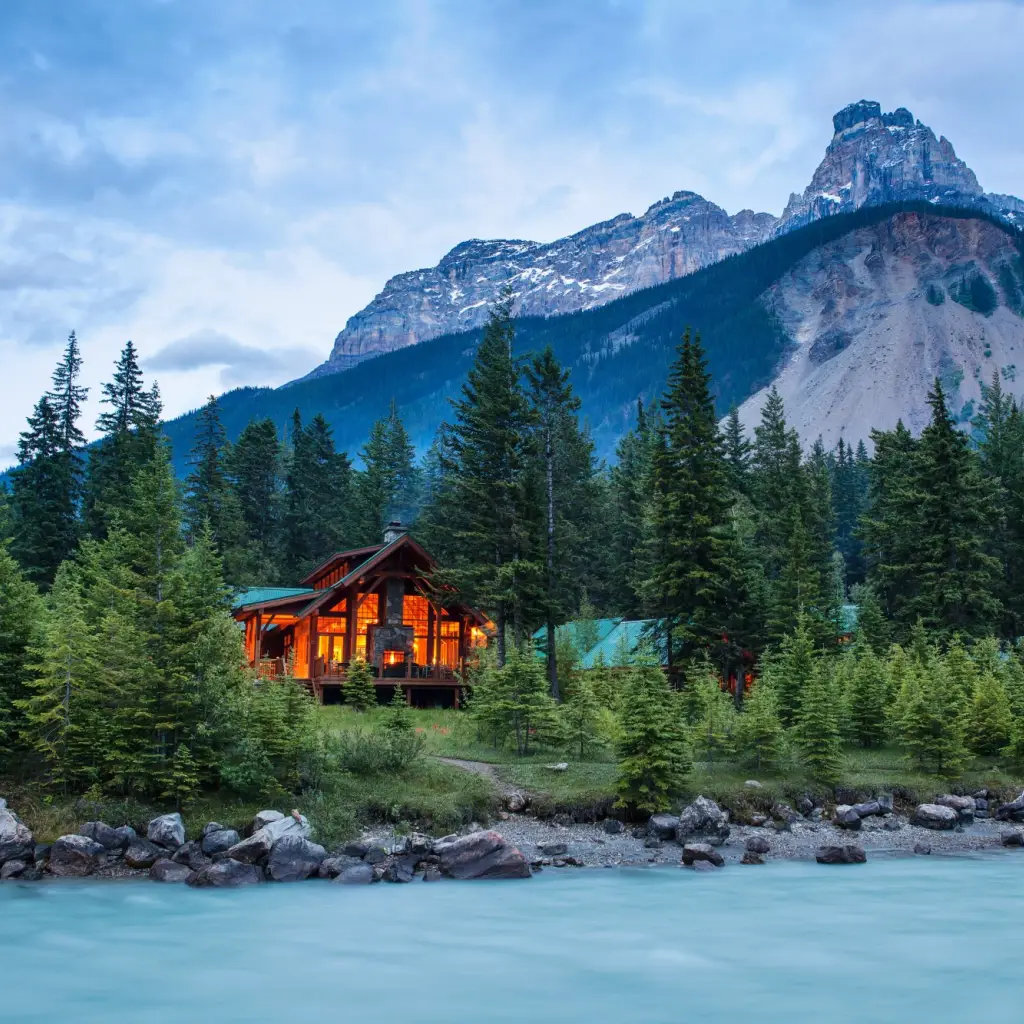 Eine gemütlich beleuchtete Berghütte aus Holz, eingebettet zwischen Fichten am Ufer eines türkisfarbenen, von Gletschern gespeisten Flusses, mit imposanten Felsgipfeln im Hintergrund im Yoho-Nationalpark, British Columbia, Kanada