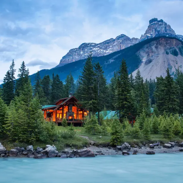 Een gezellig verlichte houten berghut, verscholen tussen de sparren aan de oever van een door gletsjers gevoede turquoise rivier, met op de achtergrond de indrukwekkende rotsachtige bergtoppen van Yoho National Park, British Columbia, Canada