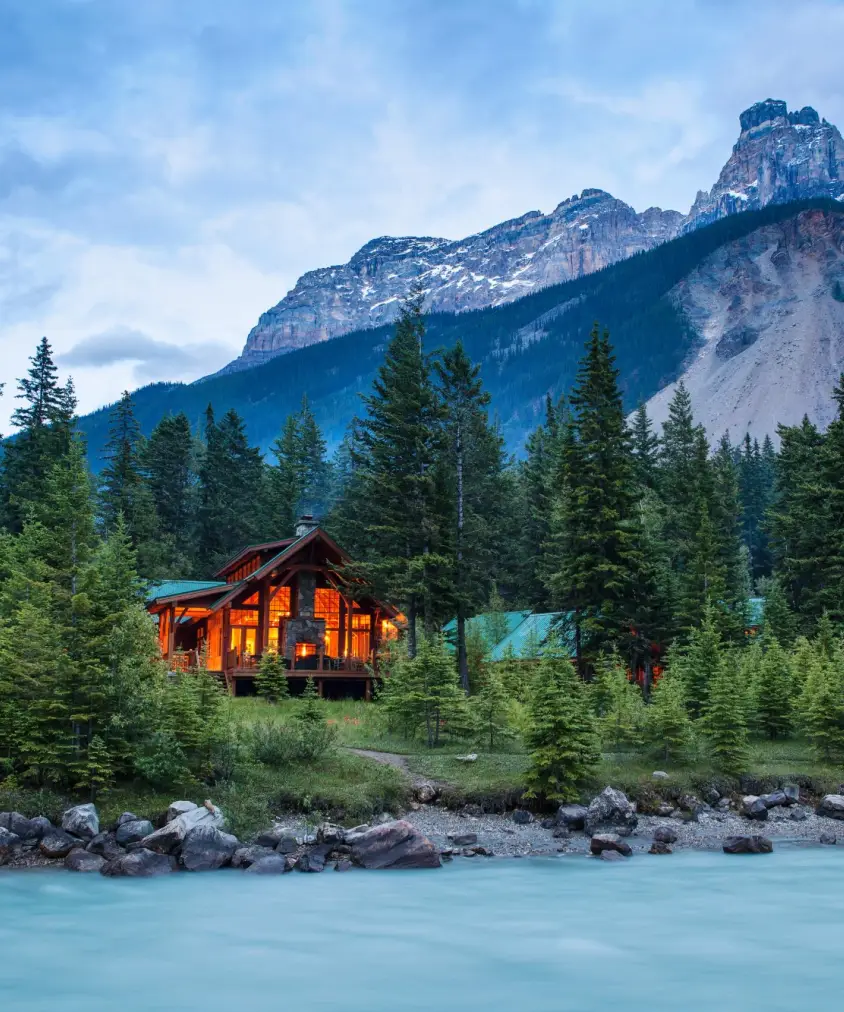 Warm-lit timber mountain lodge nestled among spruce trees on the bank of a glacier-fed turquoise river, with dramatic rocky peaks rising behind in Yoho National Park, British Columbia, Canada