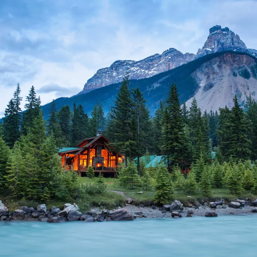 Warm-lit timber mountain lodge nestled among spruce trees on the bank of a glacier-fed turquoise river, with dramatic rocky peaks rising behind in Yoho National Park, British Columbia, Canada