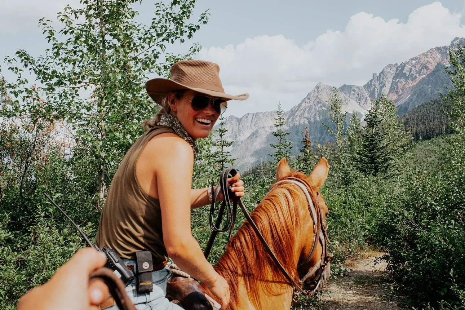 A local guide rides a horse and smiles back at the camera with mountains and trees in the background
