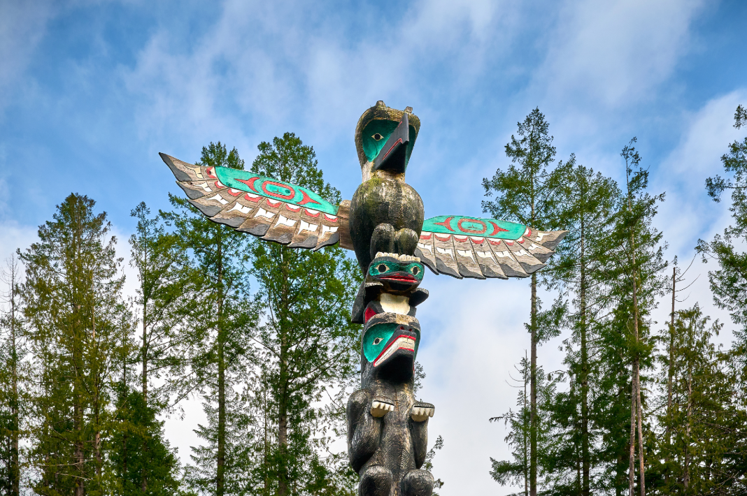 Traditioneller Totempfahl der First Nations mit einem Donnervogel und geschnitzten Figuren, die sich vor einem blauen Himmel über einem Kiefernwald in British Columbia, Kanada, erheben