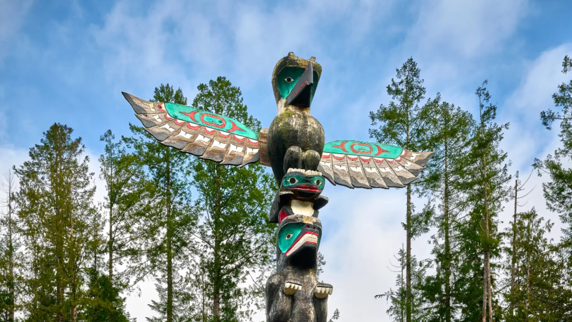 Traditioneller Totempfahl der First Nations mit einem Donnervogel und geschnitzten Figuren, die sich vor einem blauen Himmel über einem Kiefernwald in British Columbia, Kanada, erheben