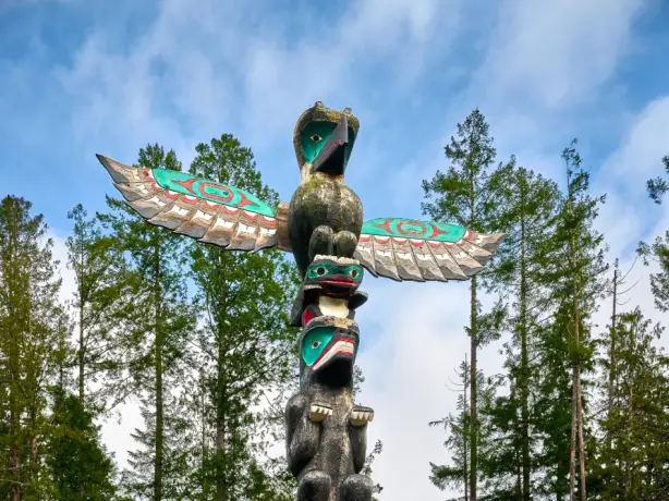 Traditional First Nations totem pole with a thunderbird and carved figures rising above a pine forest against a blue sky in British Columbia, Canada