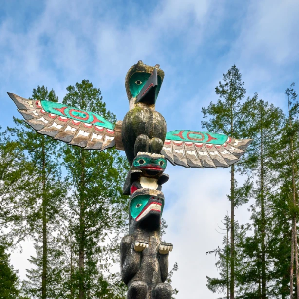 Traditioneller Totempfahl der First Nations mit einem Donnervogel und geschnitzten Figuren, die sich vor einem blauen Himmel über einem Kiefernwald in British Columbia, Kanada, erheben