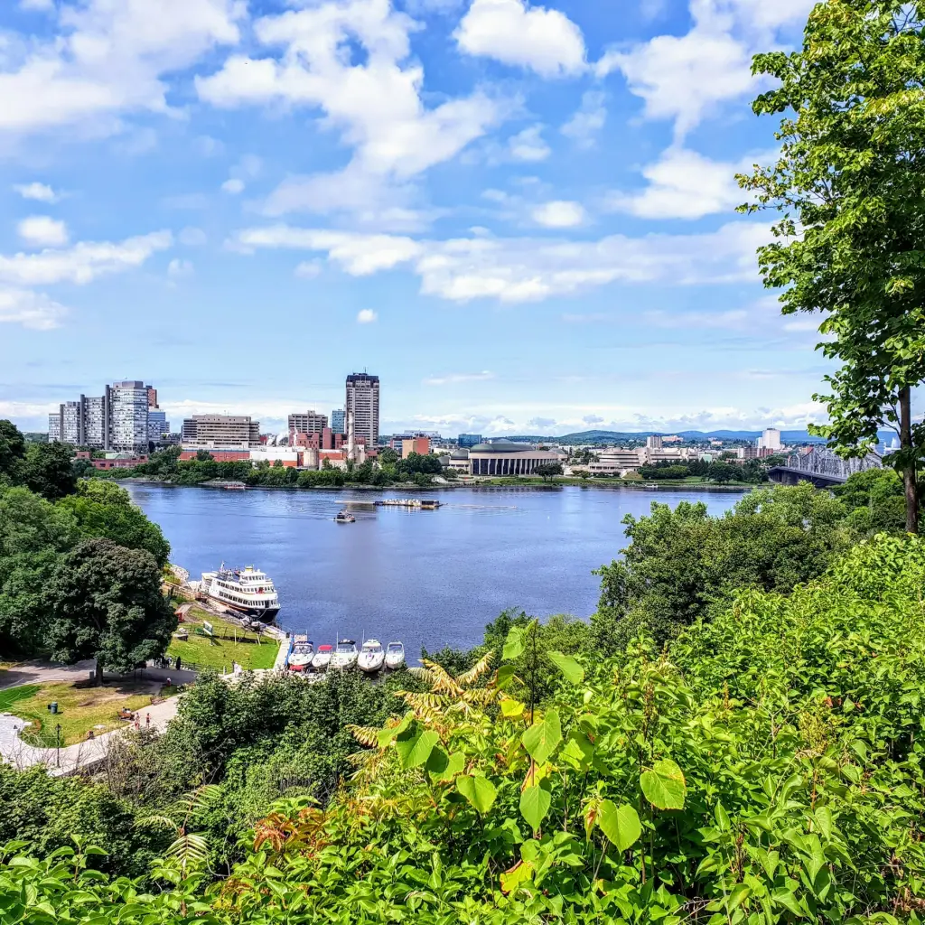Panoramablick über den Ottawa River auf die Skyline, eingerahmt von üppigem Grün und einem sonnigen Himmel
