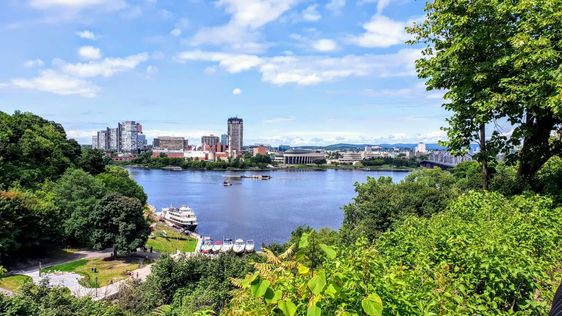 Panoramablick über den Ottawa River auf die Skyline, eingerahmt von üppigem Grün und einem sonnigen Himmel