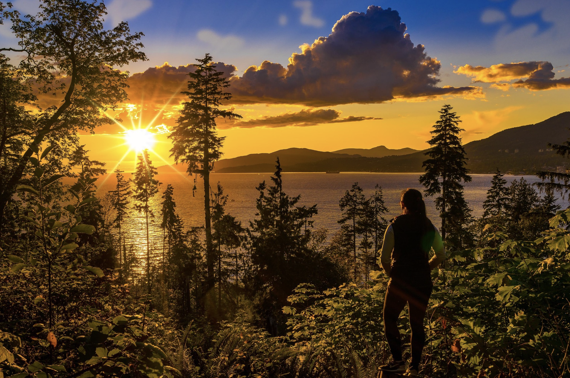 Ein Wanderer steht in einem Küstenwald und beobachtet einen leuchtend goldenen Sonnenuntergang über dem Wasser und den Berginseln von Vancouver Island, British Columbia, Kanada