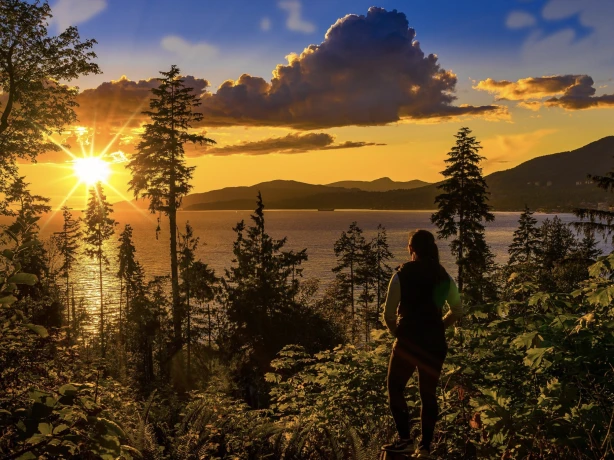 Ein Wanderer steht in einem Küstenwald und beobachtet einen leuchtend goldenen Sonnenuntergang über dem Wasser und den Berginseln von Vancouver Island, British Columbia, Kanada