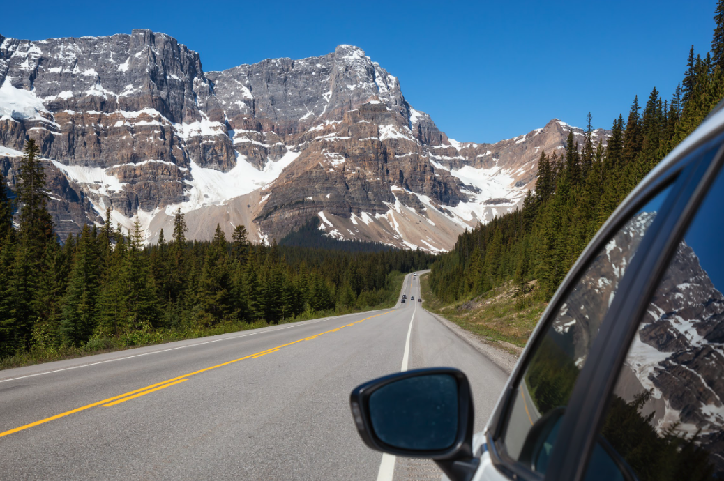 Uitzicht vanuit een autoraam tijdens een rit over de Icefields Parkway door de Canadese Rockies, met besneeuwde bergtoppen en dennenbossen die zich uitstrekken onder een helderblauwe hemel, Alberta, Canada