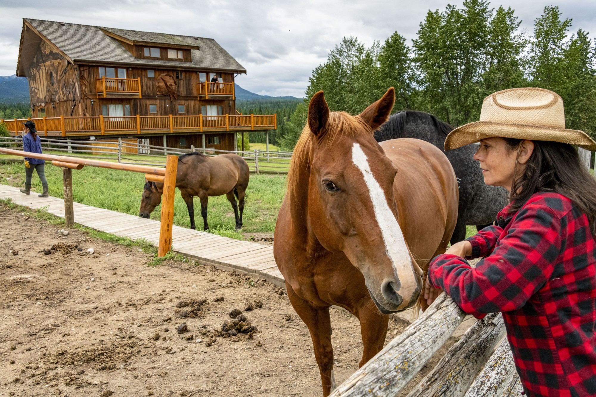 Een vrouw leunt tegen een hek naast een bruin paard, met op de achtergrond een traditionele ranch in Chilcotin, Canada