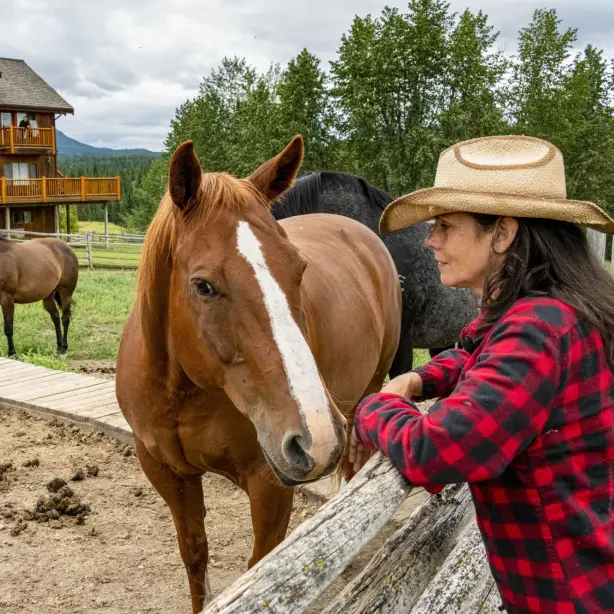 A lady leans on a fence next to a brown horse with a traditional ranch in the background in Chilcotin, Canada