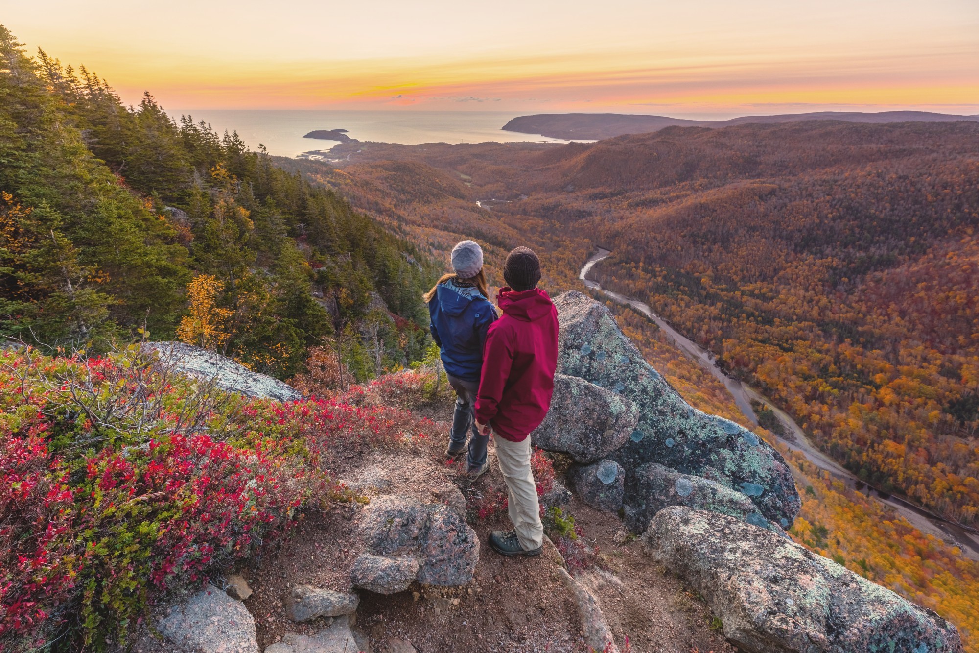Two people look out at over an autumnal forest landscape with the ocean in the distance on Cape Breton Island