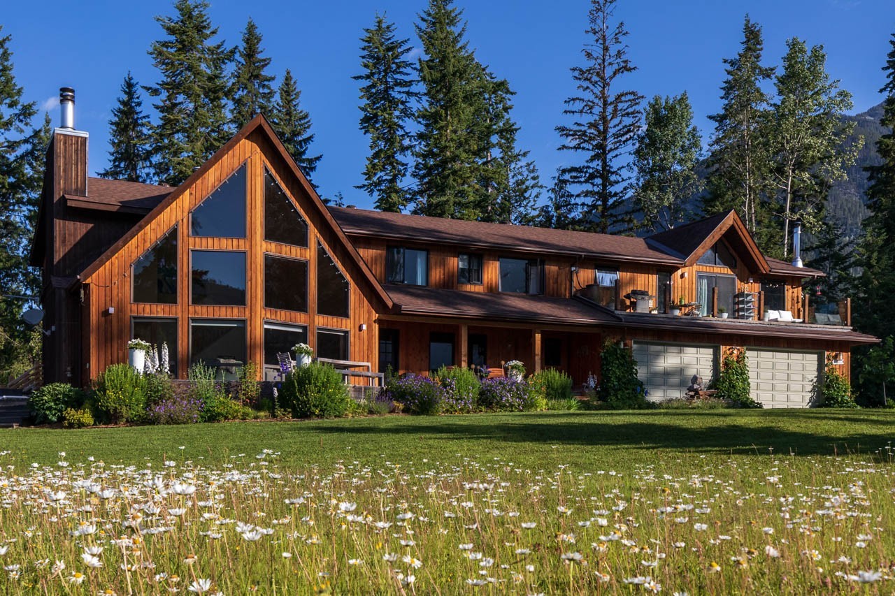 Große Holzlodge mit raumhohen Fenstern und einer Wildblumenwiese im Vordergrund, umgeben von hohen Fichten und Berggipfeln in der Nähe von Golden, British Columbia, Kanada