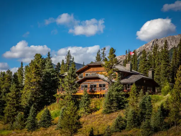 Timber mountain lodge with wraparound deck nestled among spruce trees and autumn foliage in Kananaskis Country, Alberta Canada, with rocky mountain peaks and a Canadian flag in the background