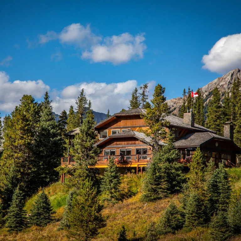 Holzlodge mit umlaufender Terrasse, eingebettet zwischen Fichten und Herbstlaub im Kananaskis Country, Alberta Kanada, mit felsigen Berggipfeln und einer kanadischen Flagge im Hintergrund