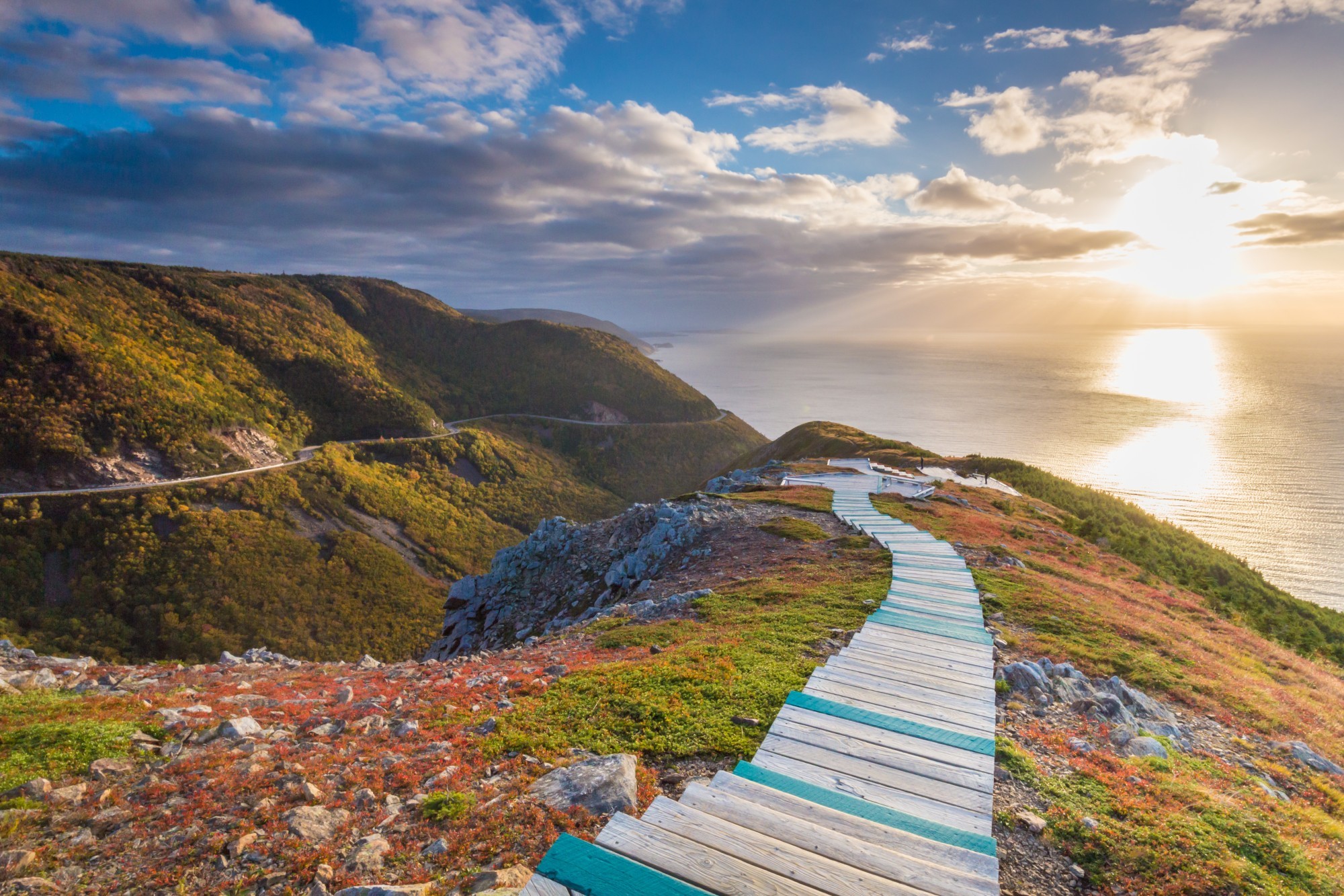 Ein Holzsteg schlängelt sich an der Seite einer steilen Klippe auf Cape Breton Island in Nova Scotia entlang, mit dem Meer und dem Sonnenuntergang im Hintergrund