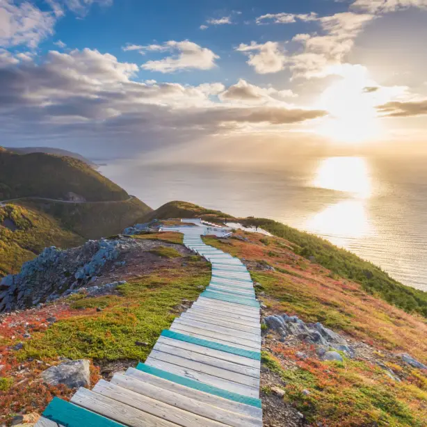 Ein Holzsteg schlängelt sich an der Seite einer steilen Klippe auf Cape Breton Island in Nova Scotia entlang, mit dem Meer und dem Sonnenuntergang im Hintergrund