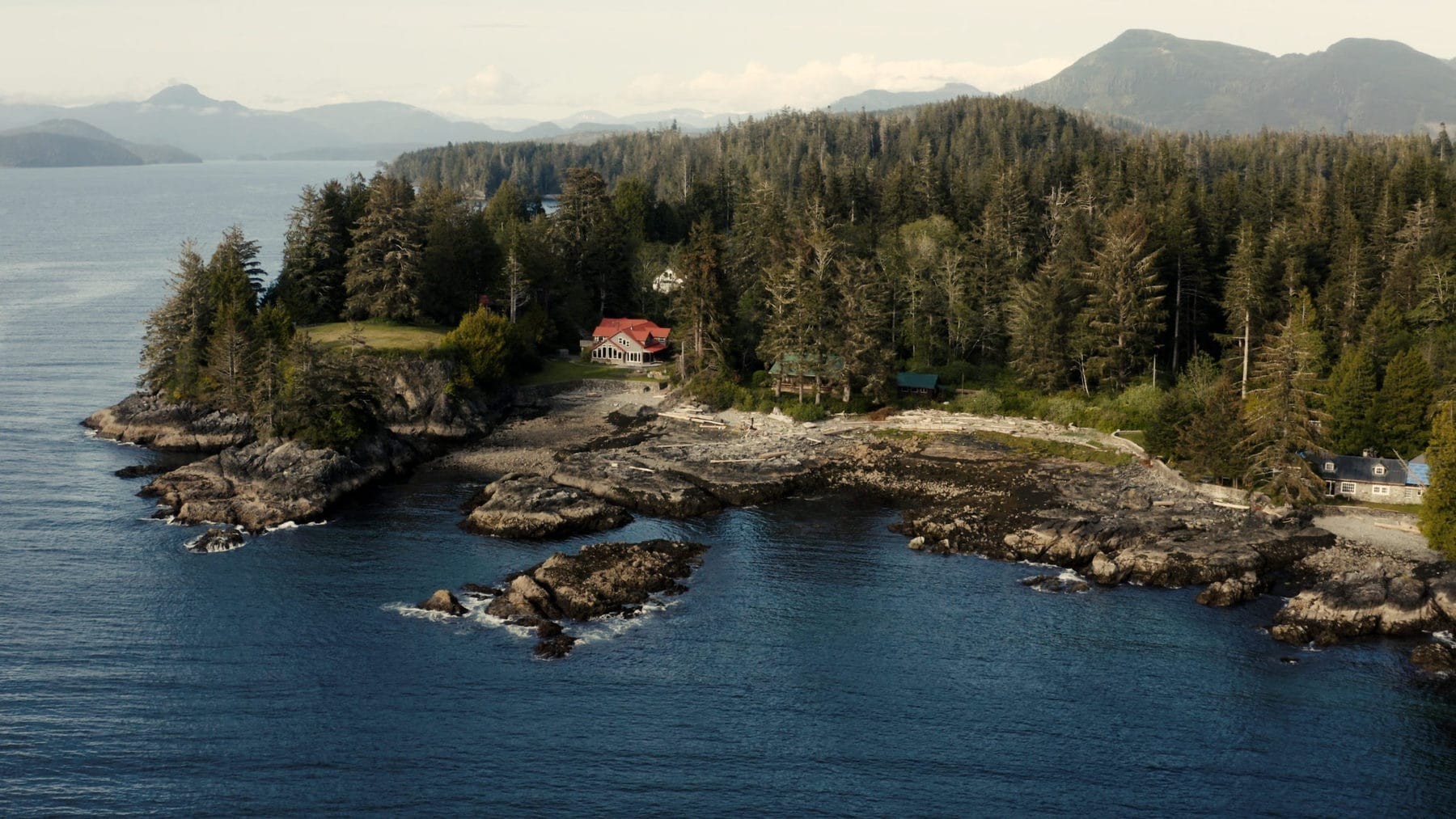 Luftaufnahme einer abgelegenen Wildnis-Lodge an einer felsigen Pazifikküste in British Columbia, Kanada, umgeben von dichtem Urwald mit Gebirgsketten über dem Wasser