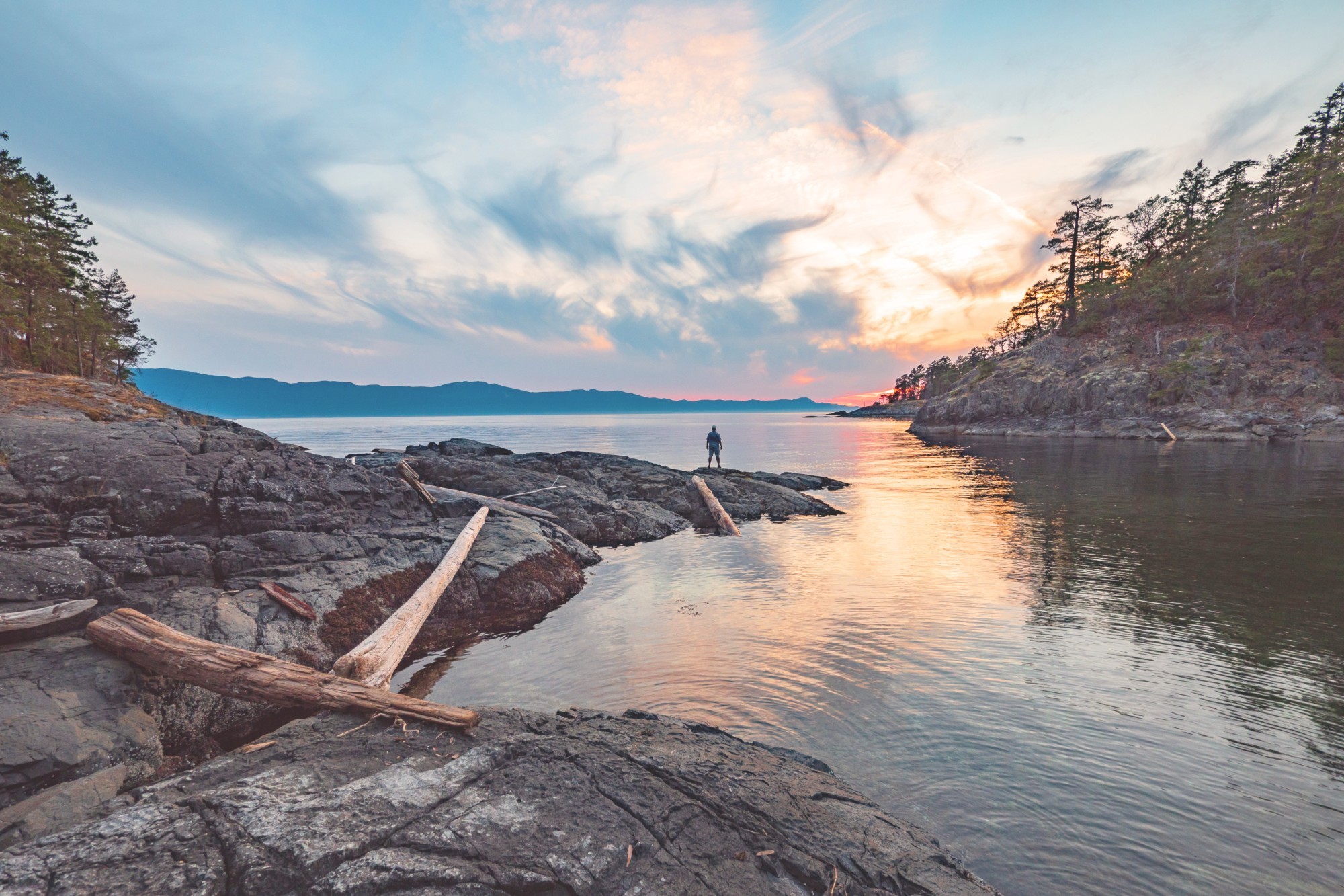 A man standing at the edge of the water looking out towards the sunset and mountains, Sunshine Coast
