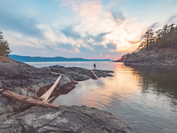 A man standing at the edge of the water looking out towards the sunset and mountains, Sunshine Coast