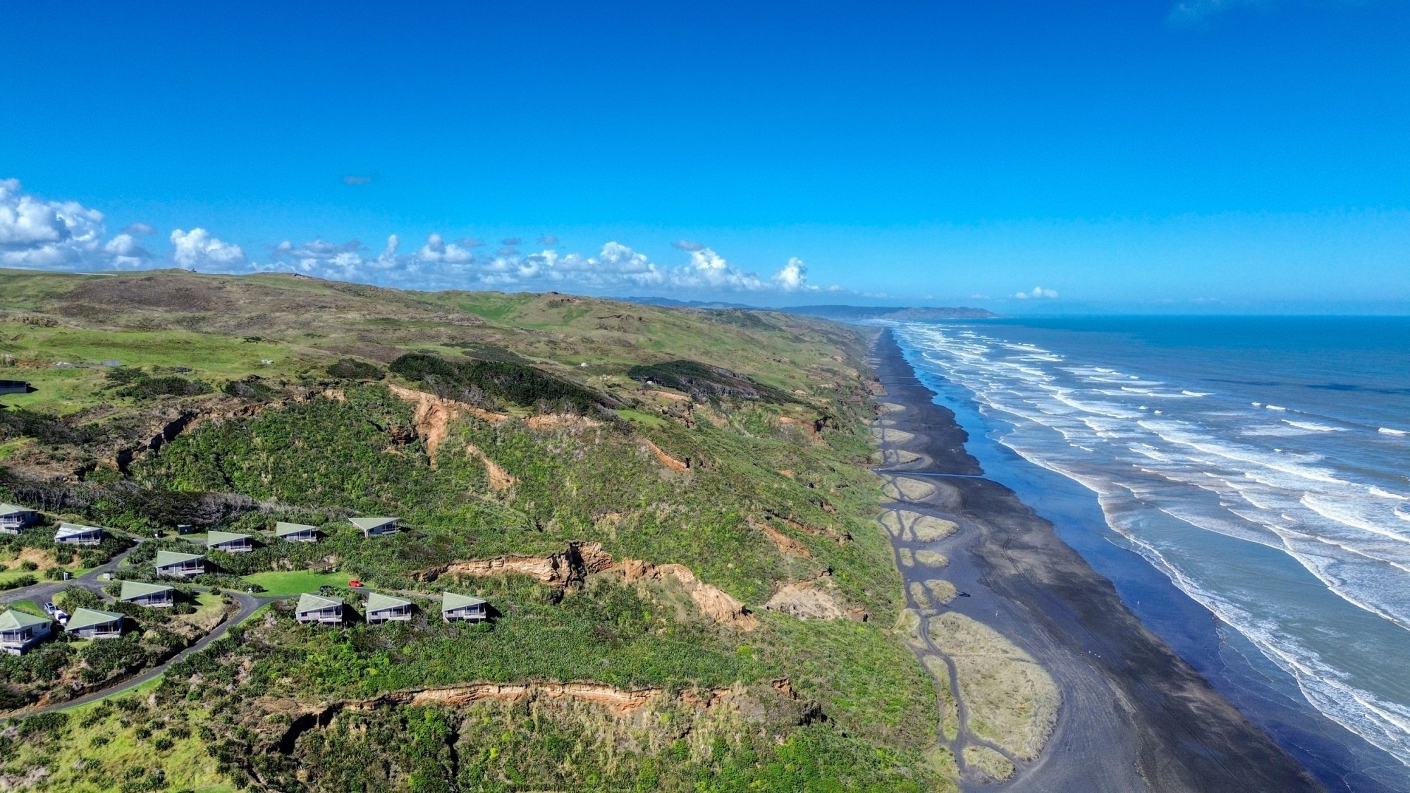 Luftaufnahme eines Klippenresorts in Neuseeland mit grün bedachten Chalets hoch über dramatischen Schwarzsandstränden und zerklüfteten Küstenklippen, die sich unter einem klaren blauen Himmel an der Küste entlangziehen