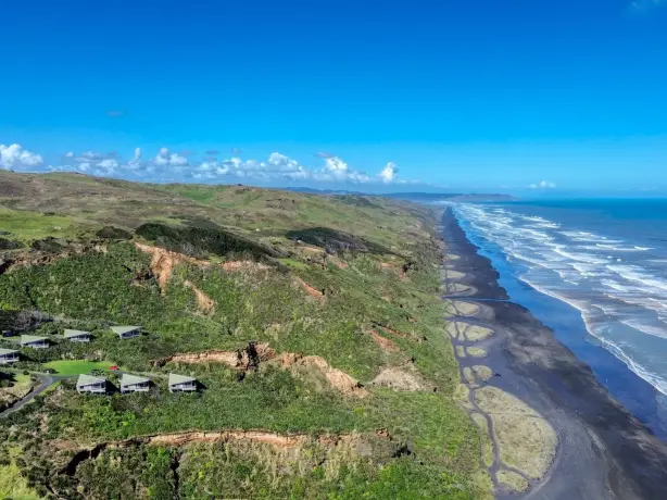 Luftaufnahme eines Klippenresorts in Neuseeland mit grün bedachten Chalets hoch über dramatischen Schwarzsandstränden und zerklüfteten Küstenklippen, die sich unter einem klaren blauen Himmel an der Küste entlangziehen