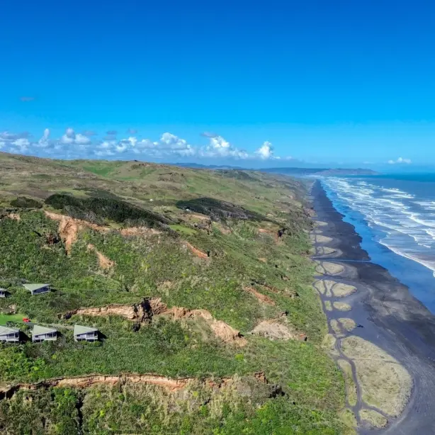 Aerial view of a clifftop resort in New Zealand with green-roofed chalets perched above dramatic black sand beaches and rugged coastal cliffs stretching along the shoreline under a clear blue sky