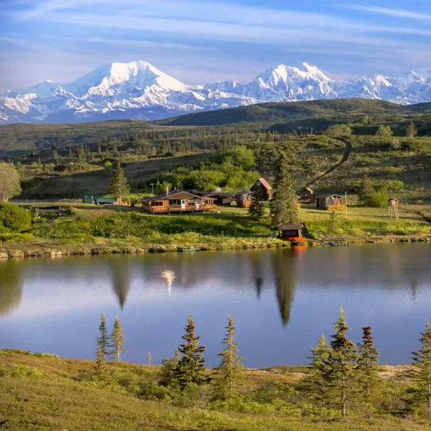 Abgelegene Wildnis-Lodge mit Holzhütten an einem stillen See in Alaska, USA, vor der atemberaubenden Kulisse der schneebedeckten Denali-Berggipfel und offener Tundralandschaft
