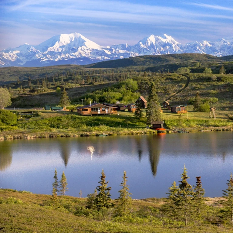 Abgelegene Wildnis-Lodge mit Holzhütten an einem stillen See in Alaska, USA, vor der atemberaubenden Kulisse der schneebedeckten Denali-Berggipfel und offener Tundralandschaft